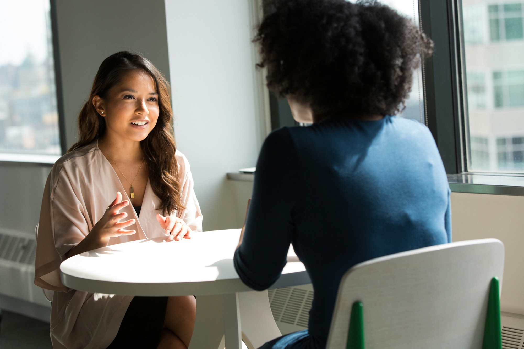 Two women in a one-on-one career conversation at a table by a sunny office window, showing the sponsor vs mentor relationship