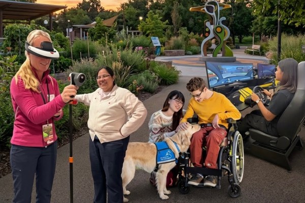 People using assistive technology in a garden at sunset: a woman wearing a headset holds a handheld device on a pole, a young man in a wheelchair pets a golden retriever service dog beside a smiling woman, and another woman sits at a driving simulator.