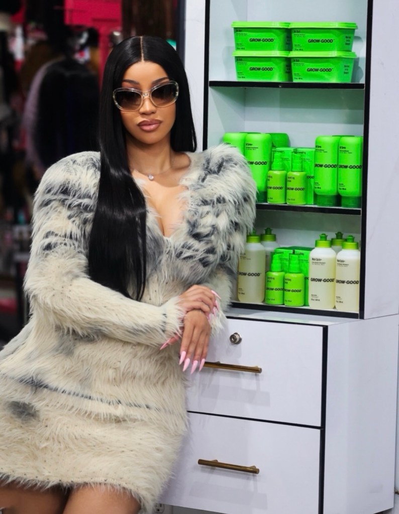 Cardi B poses beside a shelf of Grow-Good Beauty haircare products inside a beauty supply store